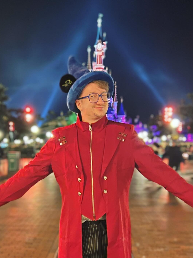 Ridgley steht vor dem Dornröschen Schloss im Disneyland Paris in einer roten Jacke mit blauem spitzen Zauberhut an denen Mickey Maus Ohren angenäht sind.