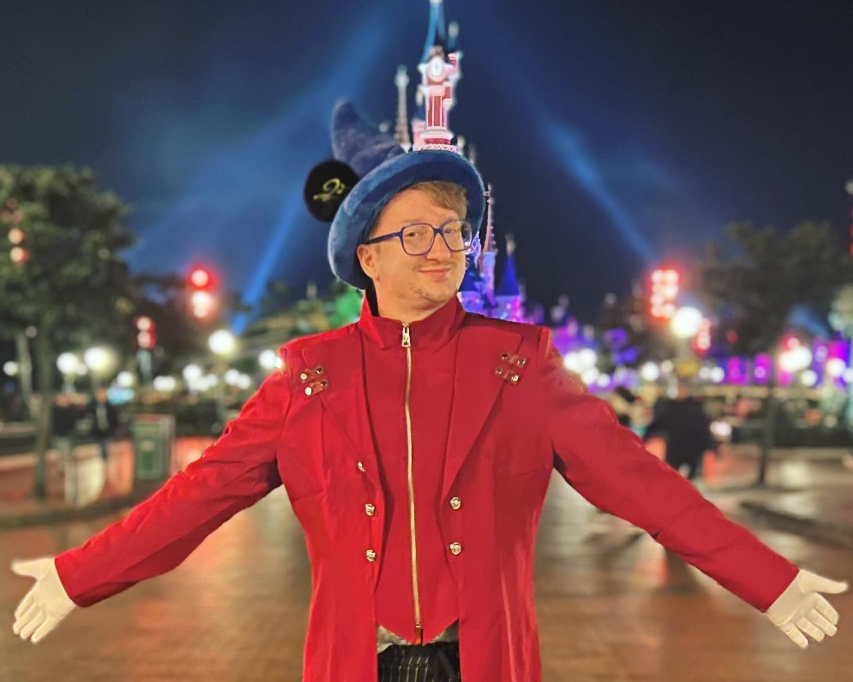 Ridgley steht vor dem Dornröschen Schloss im Disneyland Paris in einer roten Jacke mit blauem spitzen Zauberhut an denen Mickey Maus Ohren angenäht sind.